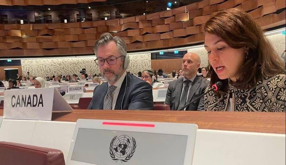 Muzna Dureid sits at a panel inside a large room with UNHCR insignia signs in front of her. A man with headphones, Peter MacDougall, sits to her left.