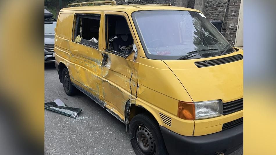 Close up of a yellow damaged van. The left side of the van is extremely damaged with dents and smashed glass across the floor.