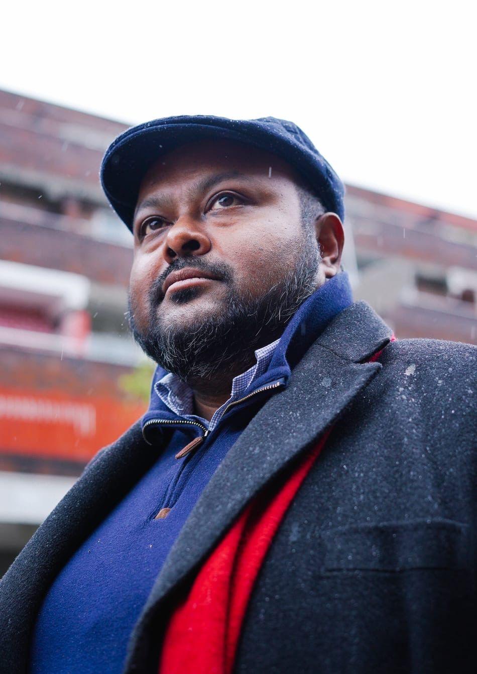 Mish Rahman, wearing a blue jumper and navy blue coat and beret with a red scarf