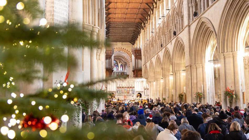 The nave of St Albans Cathedral is filled with members of the public listening to a Carols on the Hour service. There is a Christmas tree in the foreground