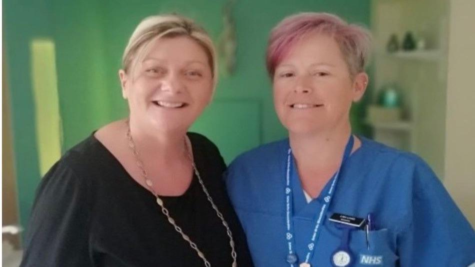 A woman in a black top stands next to a woman in blue scrubs and a name tag in a birthing room.