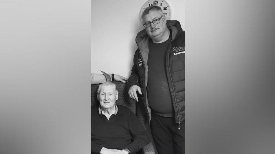 A black and white image of Ivor and Gregory McQuitty. Ivor is sitting in an armchair smiling as Gregory stands leaning onto the armchair also smiling. Ivor has short light hair and is wearing a jumper. Gregory has short light hair and a moustache, he is wearing glasses and a jumper with a coat over it.