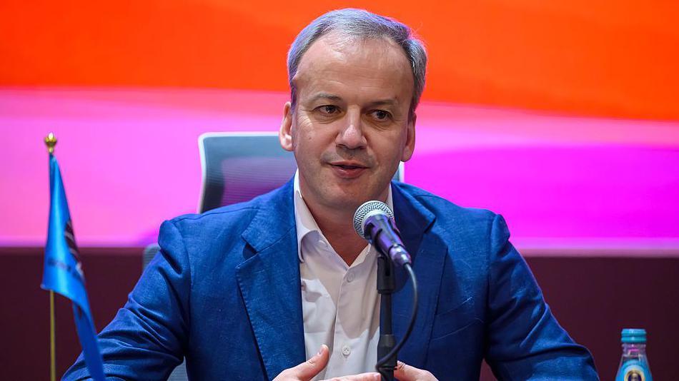 Arkady Dvorkovich is speaking into a microphone. He is sat down. He is wearing a blue blazer with a white shirt underneath. 
