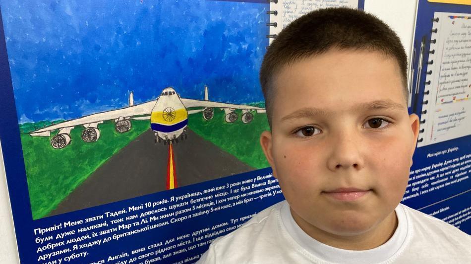 A 10 year old boy with brown hair stands in front of his contribution to the exhibition - a drawing of the Antonov An-225 plane - a symbol of national pride for Ukraine, the cargo plane's livery is white with yellow and blue strips across the centre.