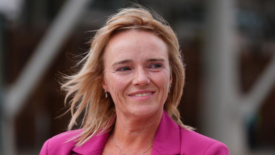 Nurse Sandie Peggie has short blonde hair and wears a pink blazer and smiles looking off to the side.