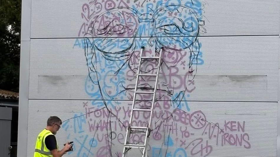 An outline of a mural, showing words and a face, with a man in a high-vis jacket, with paints in his hands, and a metal ladder in the middle. 