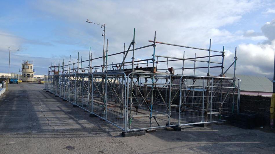 Scaffolding poles have been connected to create a long tall structure on a tarmacked pier. Barriers have been est up around it. The sky is blue.