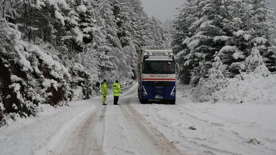 Police officers wearing bright clothing stand next to the lorry. There is snow all over the road and on the trees next to the road.