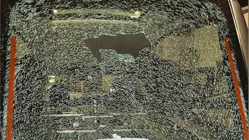 The back window of a hearse. It has been damaged. There is a large hole midway up the window. It is surrounded by cracked glass. 