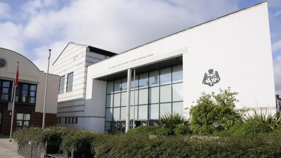 A modern courthouse building with a large glass entrance, and the Isle of Man government crest on the well. Green bushes and wooden benches are in the foreground. 