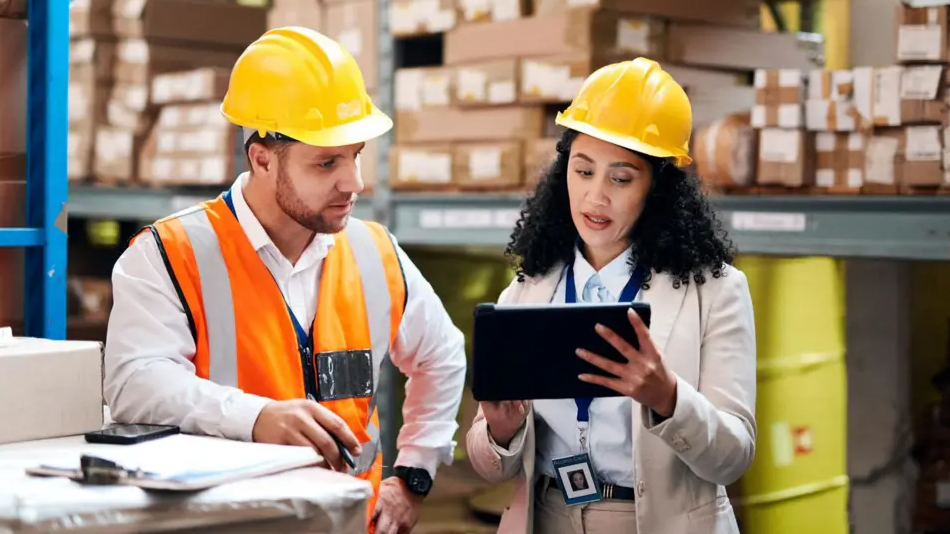 A man and a woman are talking inside a factory. They both have yellow safety helmets on and the woman is holding a small computer. The man is also wearing a fluorescent jacket.