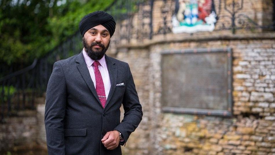 Mr Sohal is wearing a suit with a pink short and darker pink tie. He wears a turban and is stood outside by a brick wall.