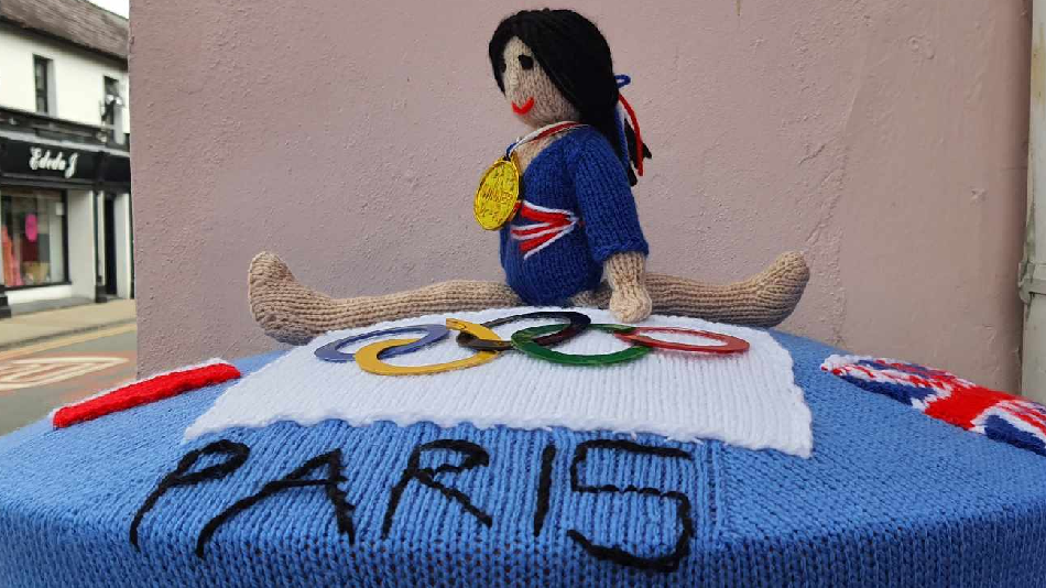 Sue Turner's knitted postbox topper of a GB gymnast with a gold medal around her neck on a blue mat with Paris and the five Olympic rings