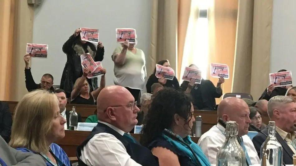 A row of councillors seated on a bench stare forward while behind them a group of protesters in a council hall's public gallery stand up holding pieces of paper calling for a child sex abuse national inquiry. 