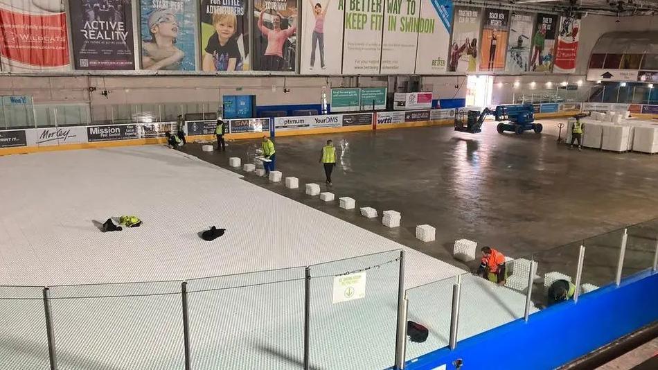 An ice rink with half of its floor removed. There are several workers wearing high vis vests