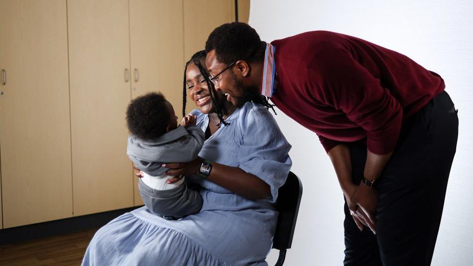 Lorna Mulungi with her husband, Yassir, and their baby. Lorna is seated and has long black hair. She is wearing a light blue dress and holding the child. Yassir has short dark hair and is wearing a dark red jumper and black trousers. He is leaning in as the pair smile towards their baby.