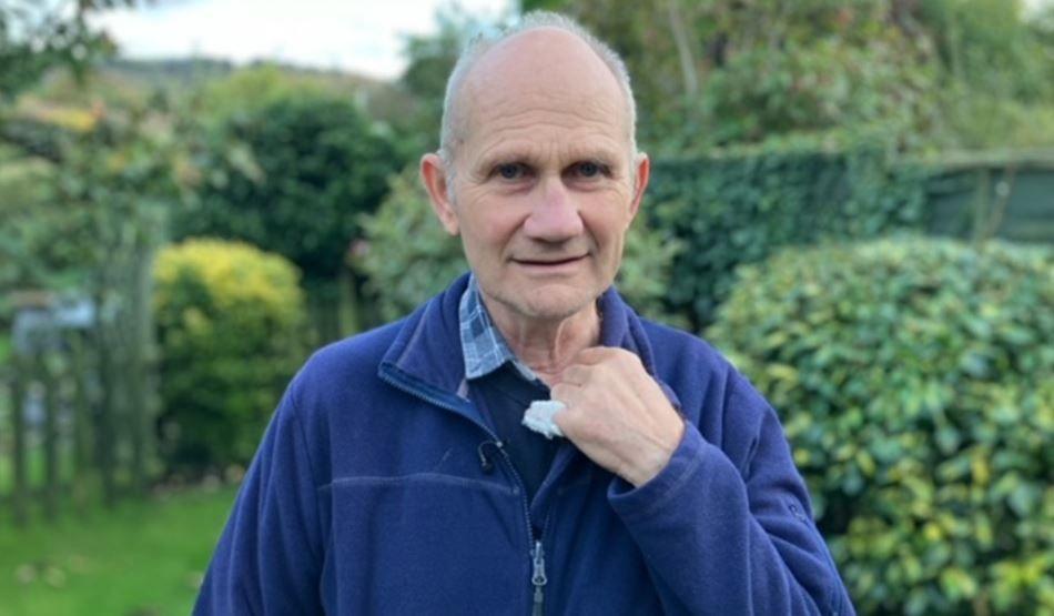 A man with grey hair looks at the camera. He is standing in a garden with green hedges in the background. He is wearing a navy blue fleece over a blue humper and blue checked shirt.