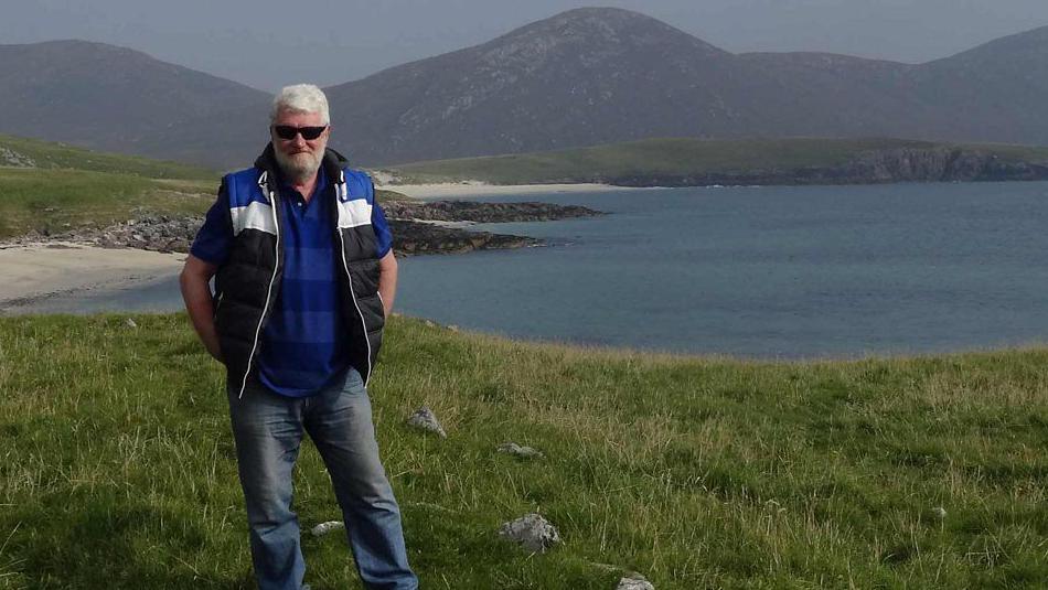 Iain stands in an island landscape with a loch and hills behind him. He is wearing sunglasses and has his hands in his pockets.