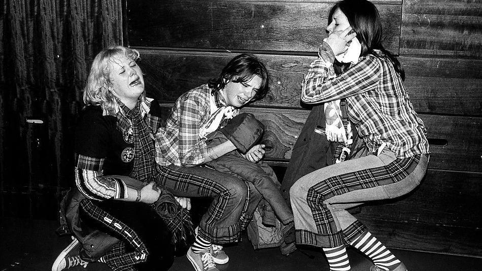 In this black and white shot, three ecstatic fans are brought to their knees with excitement at a Bay City Rollers concert. The first is openly sobbing, kneeling on the floor in her tartan cropped bell bottoms. Her friend, in a similar outfit, cries as she leans against a wall. The third is bent down, holding her head in her hands.