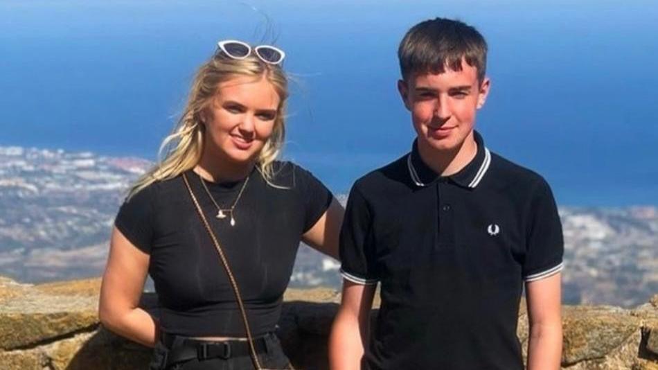 A young woman with long blonde hair dressed in black stood beside a young man with short brown hair wearing a black Fred Perry polo top. They are looking into the camera and smiling while stood with their backs facing what appears to be a viewing point above a coastline