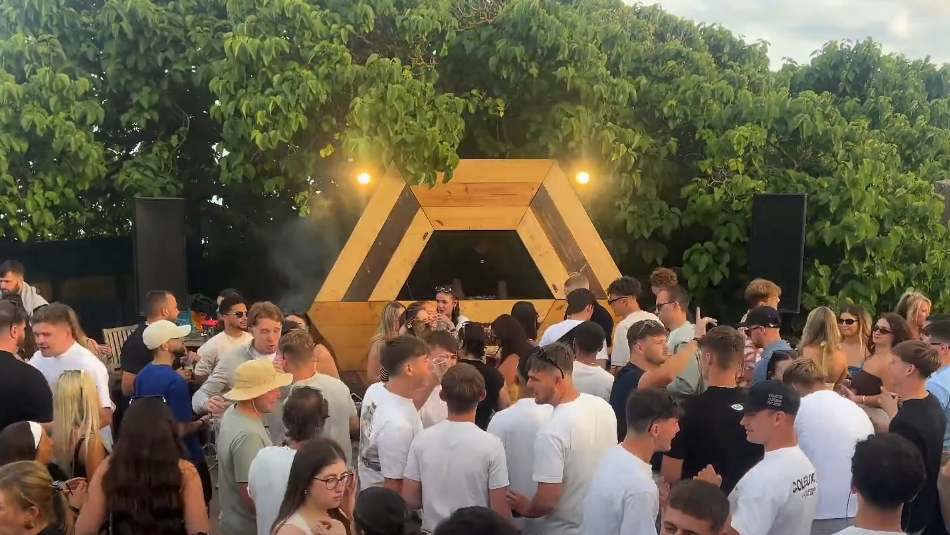 A crowd of people standing in front of a hexagon shaped DJ stage, with trees in the background and grey skies.