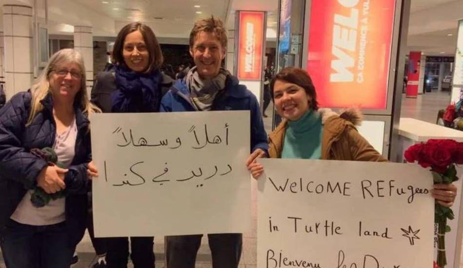 Four people stand in an airport holding signs that read "welcome refugees to Turtle Island" in English, French and Arabic.