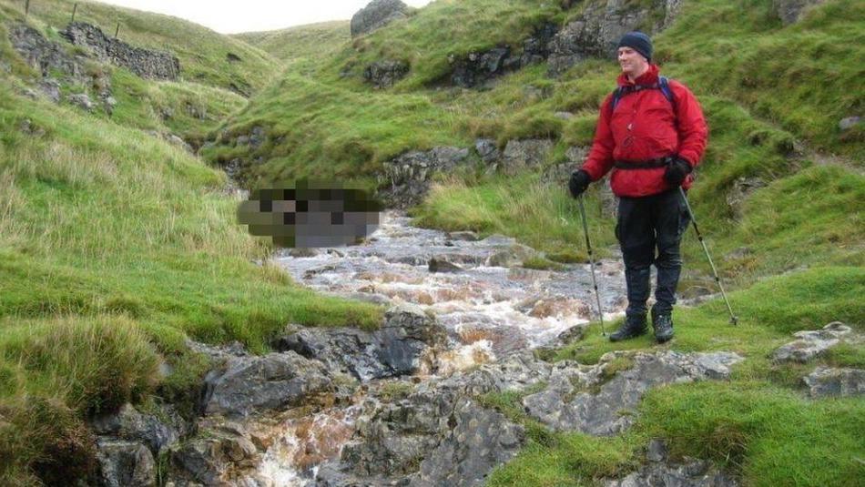 A man posing next to a rocky stream running through hills in the Yorkshire Dales. The man is wearing a red jacket, black trousers, a hat and gloves and is holding walking sticks.