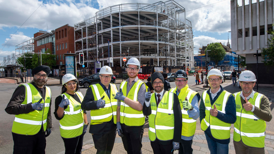 A group of eight men and women in hi-viz jackets and safety helmets stand in front of an under construction building while giving the thumbs up