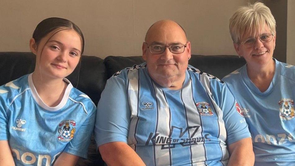 A man sits on a sofa next to a woman and a girl in a living room. All three are wearing Coventry City FC t-shirts. A dog is also sat on the floor.