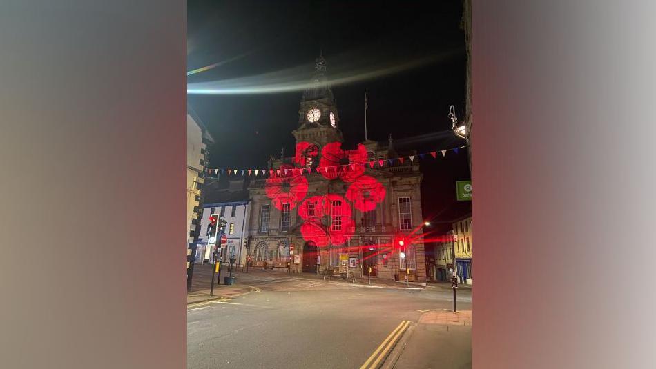 Kendal Town Hall is an impressive stone building with a central clock tower, arch windows on the ground floor and high rectangular ones on the first floor. It sits on a road junction and is attached to an old white fronted building. It is shown at night with a circle of five poppies beamed on to its front.