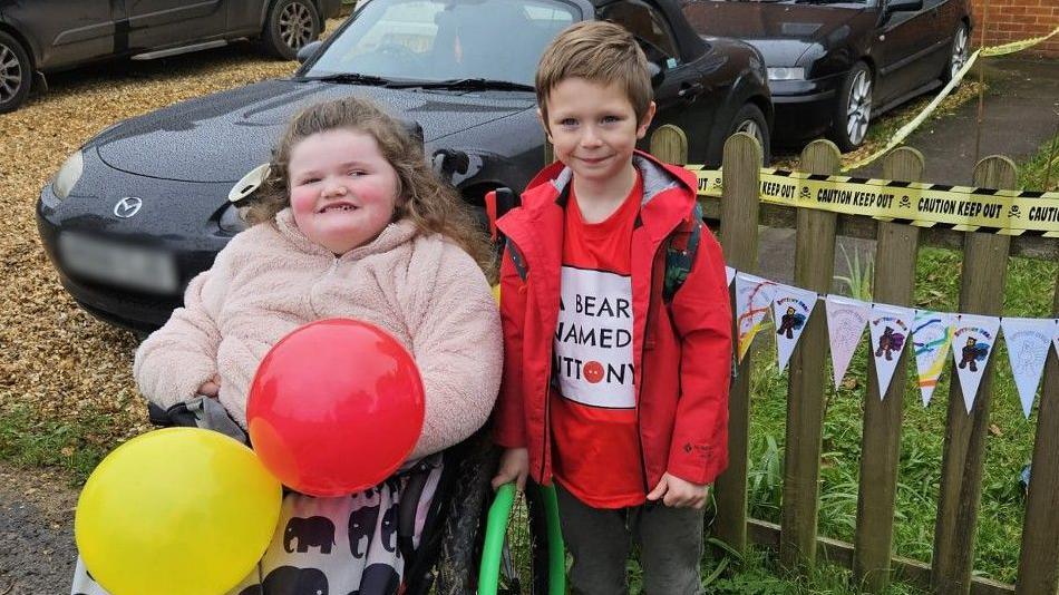 Riley, who is wearing a red coat and t-shirt is standing in front of a fence alongside his friend Ellie. Ellie is holding a yellow and red balloon and is wearing a cream fluffy jacket.