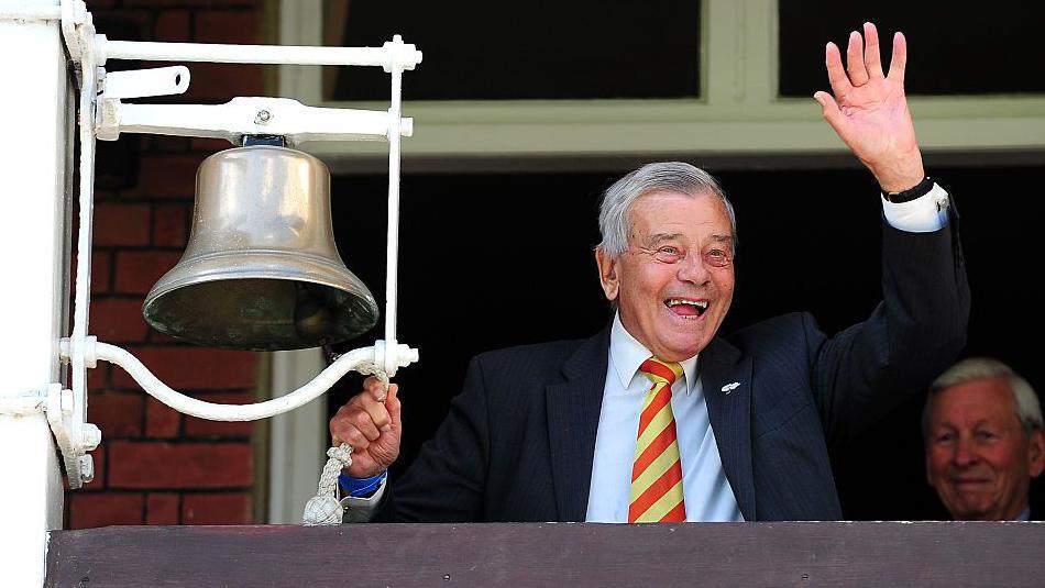Retired international cricket umpire Dickie Bird rings the five minute bell prior to the start of play during day one of the 1st Investec Test match between England and New Zealand at Lord's Cricket Ground. Bird wears a dark jacket and a red and yellow striped tie and is waving with his left hand as his right pulls the bell.