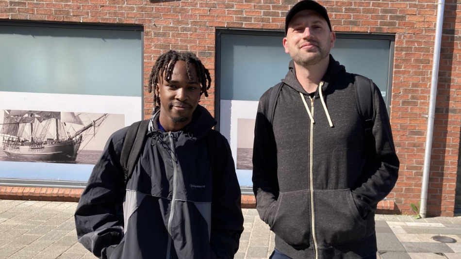Two men stand outside a brick building with large windows displaying images of historical ships. The man on the left wears a dark jacket and carries a backpack; the other wears a black hoodie and cap. 