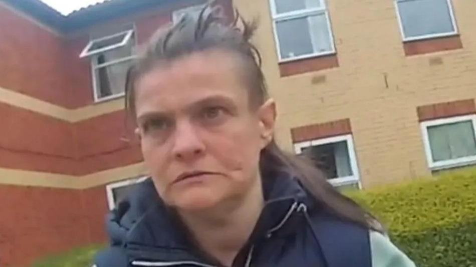 Claire Driver, wearing a navy hooded gilet, standing in front of a block of flats  