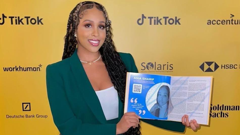 Asia Sharif stands in front of an even board, holding open a book which tells her story about her journey with cancer and the work she is doing to raise awareness of the need for stem cell donors.