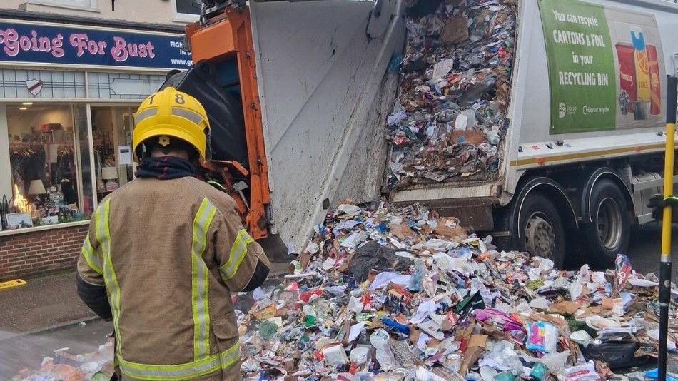 The contents of a waste collection truck spilled across a street. A worker wearing a helmet is standing at the rear of the truck.