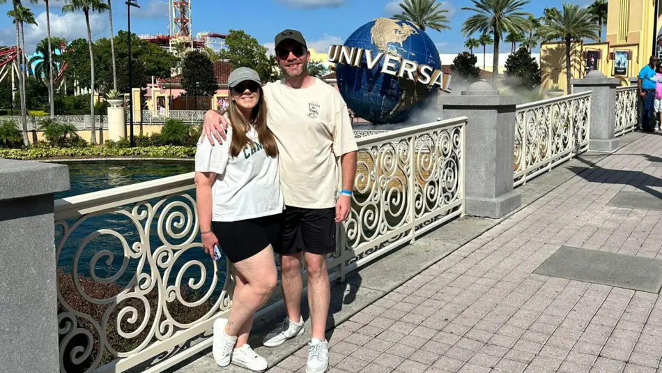 A woman with a white t-shirt, black shorts, white trainers, a grey baseball cap and sunglasses standing next to a man wearing a cream t-shirt, black shorts, white trainers, a black baseball cap and sunglasses against white railings, with the Universal Orlando resort in Florida pictured the background.