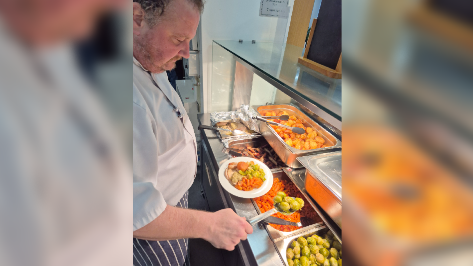 A man in a striped apron and white shirt serves a roast dinner from a serving hatch. 