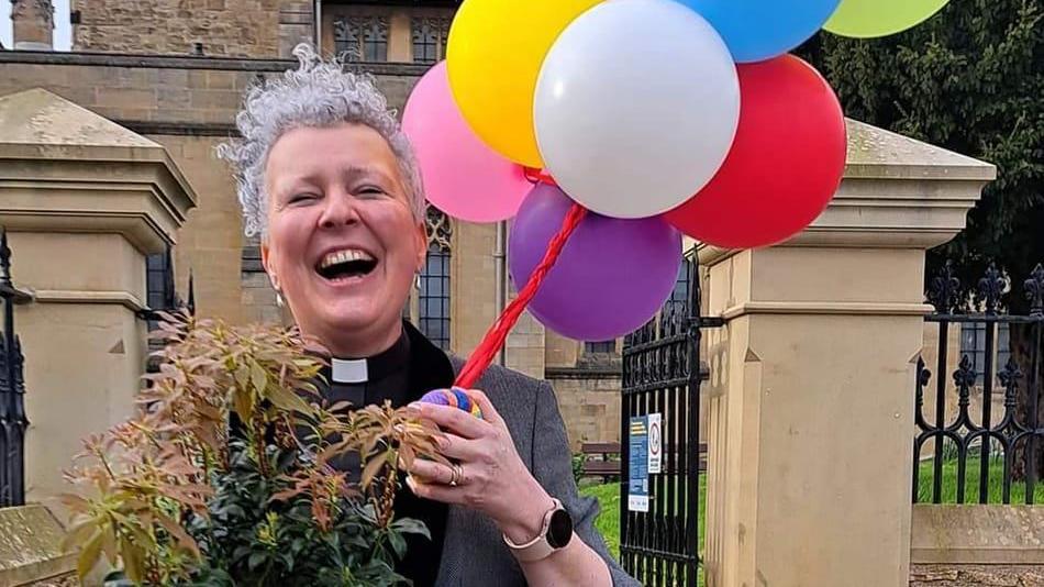 Clare MacLaren, wearing a clerical collar and laughing into the camera. She has short grey hair, and is pictured holding a plant and a colourful buunch of balloons.