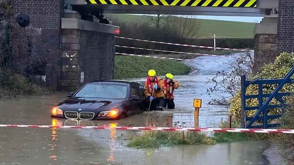 A black car stuck in floodwater on a road. The murky brown water is nearly at its headlights. Two firefighters are standing next to the car. The road has been taped off.