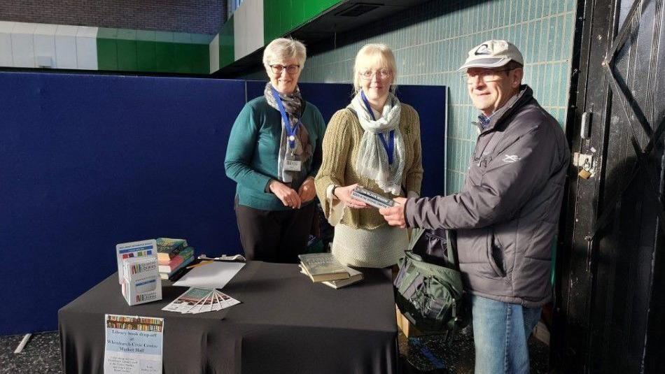 Three people stand at a table with books in their hands as a pop-up library service is provided. There are leaflets on the table in a stand and laid out on the tabletop, and there are more books on the surface waiting to be collected. The table has been placed near a screen in a hall, near a tiled wall. The two women are wearing jumpers and scarves and a man is wearing an anorak and a peaked cap.