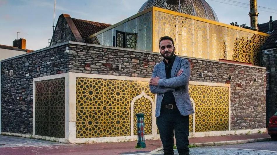 A man stands in from of a mosque with his arms folded. He wears a grey jacket and a black shirt and trousers. The most has a dome and is covered in black and gold tiles. 