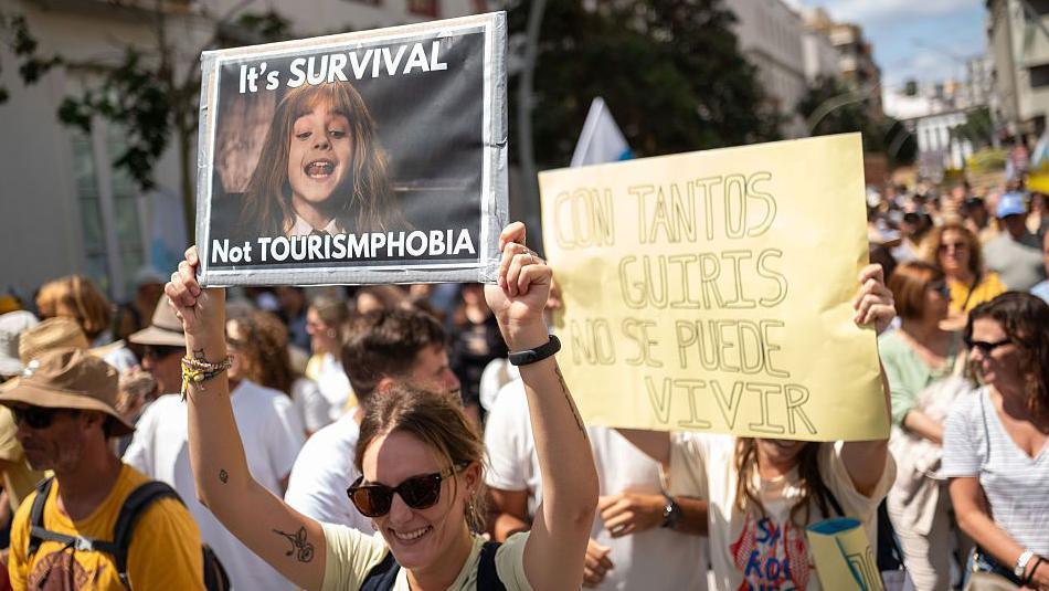 A woman wearing sunglasses holds up a sign that reads: It's Survival, Not Tourismphobia