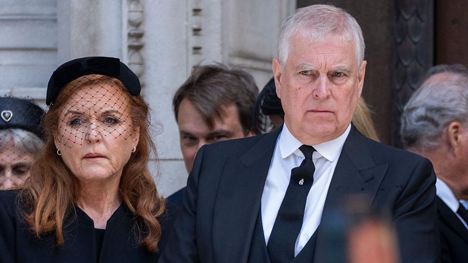 Sarah Ferguson is standing on the left of the image wearing a black outfit with a small black hat and veil, Prince Andrew wearing a black suit and tie stands beside her