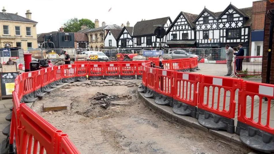 Orange barriers surrounding a road where resurfacing works is taking place. Black and white timbered houses and shops and stone buildings, as well as vans, cars and pedestrians are in the background.