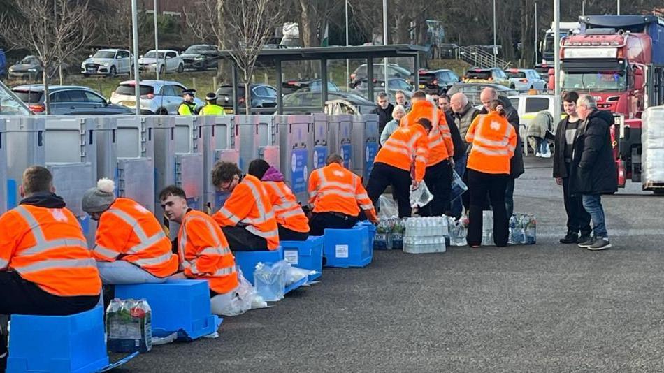 Picture of staff members in orange hi-viz jackets sat on blue crates and surrounded by bottles of water at the water station in Zip World Conwy, with queue of cars in background