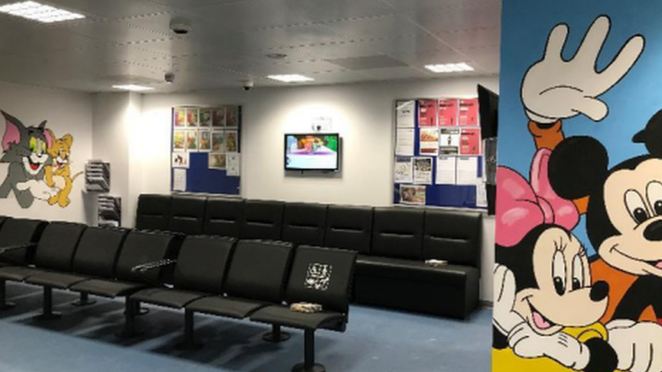 A waiting room with a blank white wall where the mural has been painted over, with the previous Mickey Mouse mural shown on the right hand side.