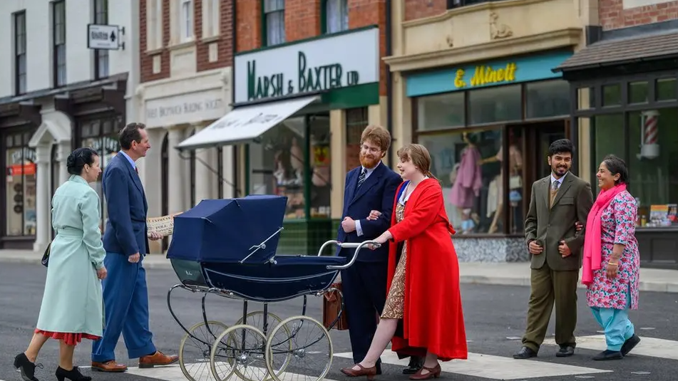 Men and women dressed in 1950s attire cross a road at the museum. One couple is pushing a pram with a South Asian couple walking behind them and another couple walking towards them. 