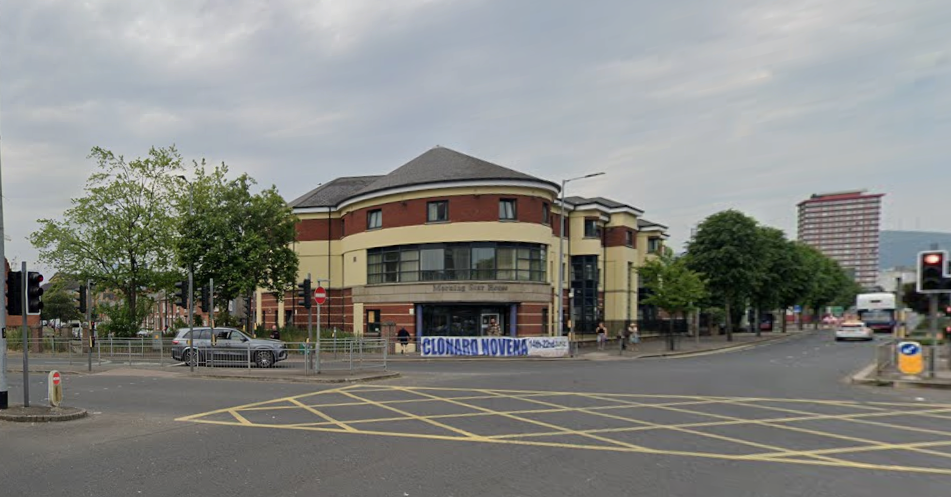 A street view of a yellow box junction. A grey jeep can be seen behind a pedestrian island with railing and traffic lights. There is a brick and yellow building in the centre behind the junction. Trees can be seen along the roadside.