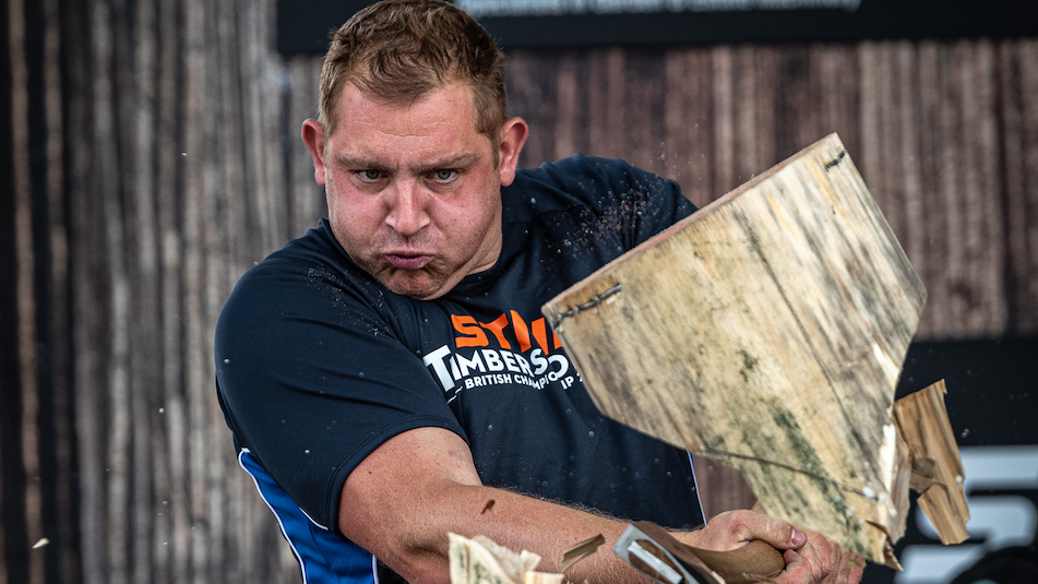 A man wearing a black, dark blue and white T-shirt with an orange and white logo on it is chopping a log of wood. There is a wooden wall behind him.
